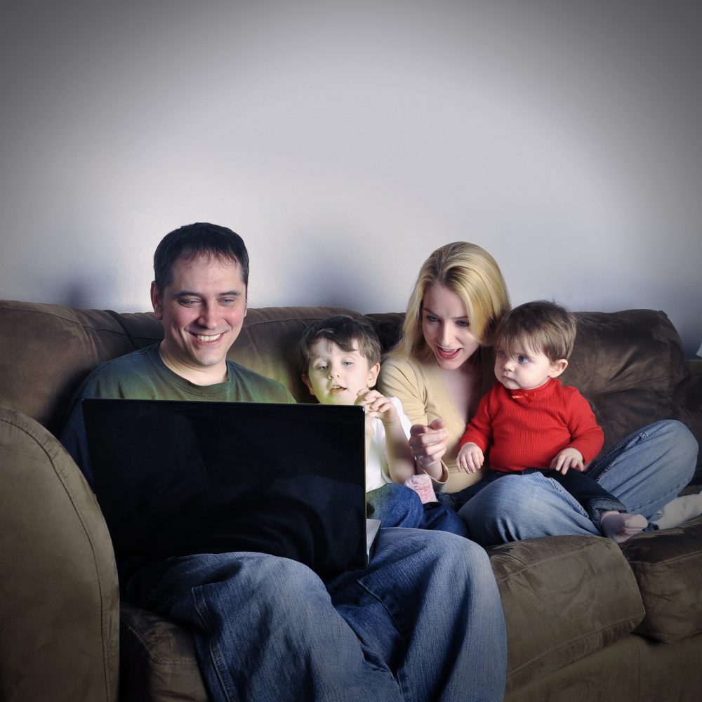A modern family is sitting on a couch in their home watching a computer internet screen and look happy and excited. Use it for a media technology concept.